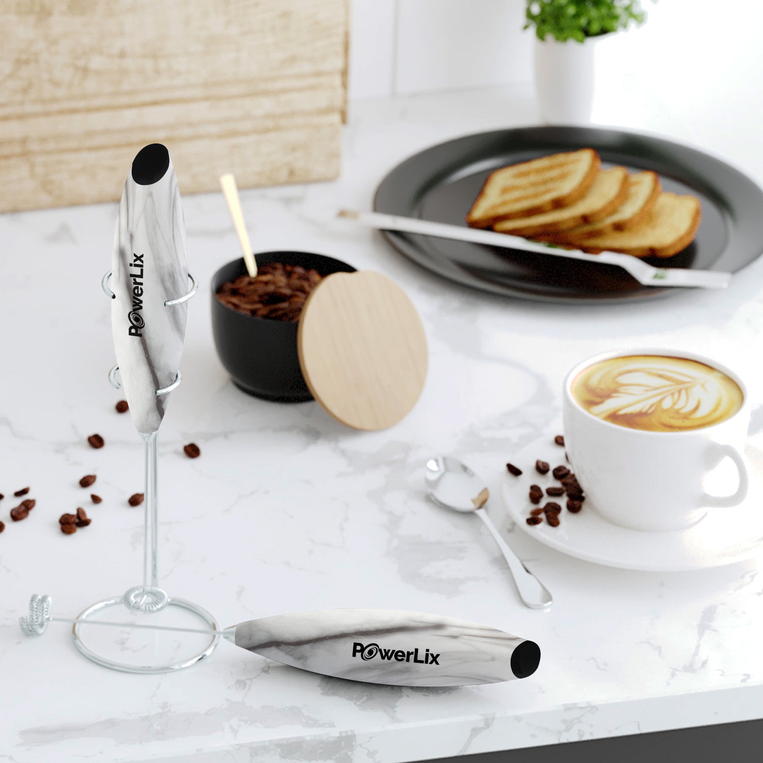 Powerlix milk frother in marble on its stand with another on its side on a counter. Ther is a coffee cup behind it with a plate of toast and a container with coffee beans.